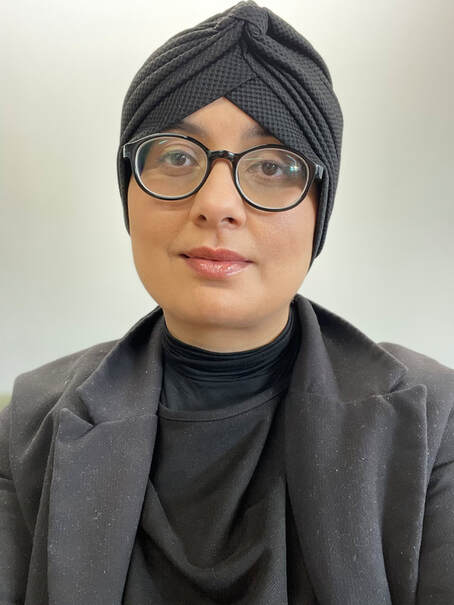 Sahera Khan is a young Musilm woman wearing greay top and jacket, along with dark rimmed glasses. Sahera is smiling to camera and she is wearing an Islamic head covering to cover her hair. 