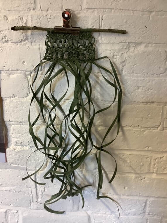 A bulldog clip against a white brick wall holds a 15 inch long thin branch onto which Steph has used balck 1cm strands to create traditional macrame in a non-traditional way. 