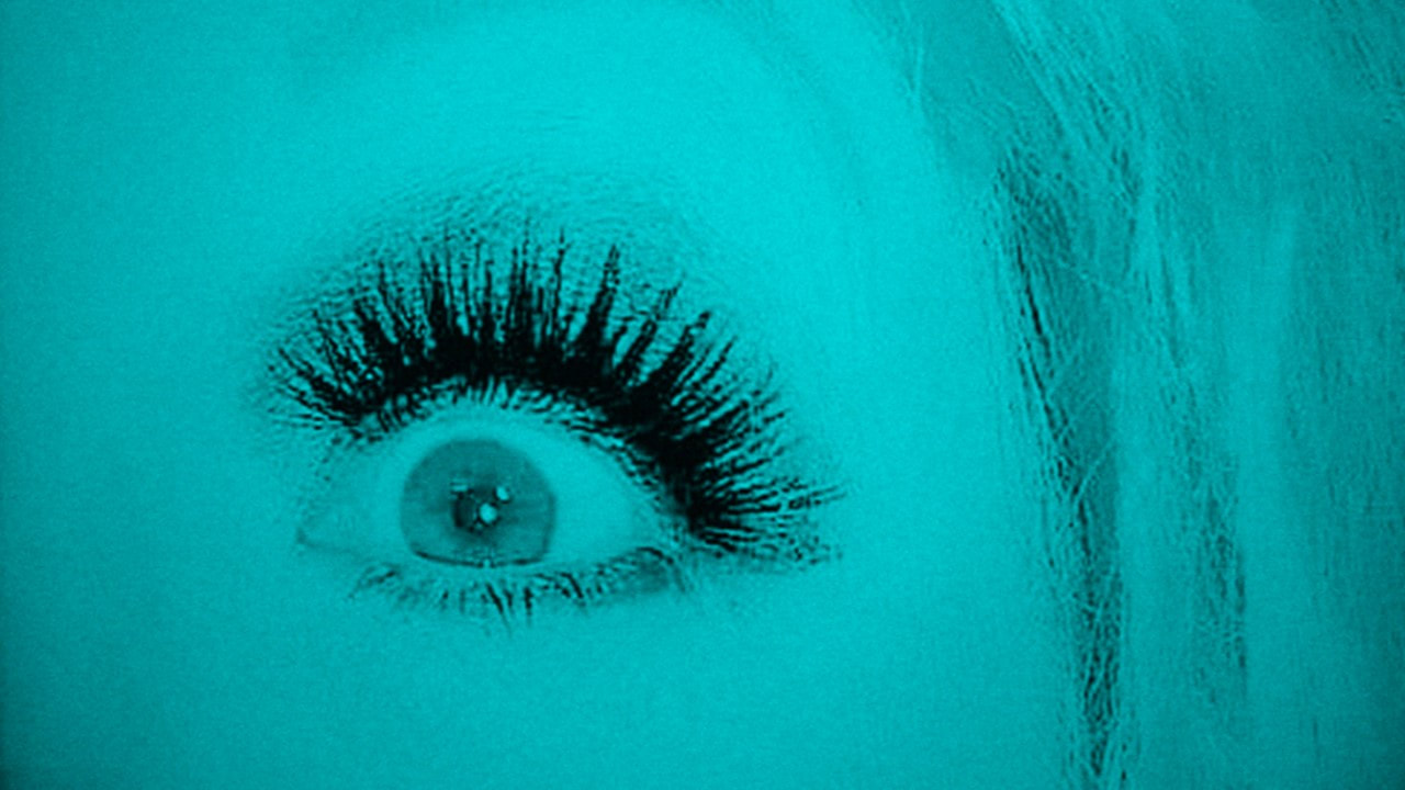 A turquoise image of a single eye with long thick eyelashes. A large teal coloured wig is visible just to the right or the persons head and image. This is a promotional photo from the Wrong Woman Discussions. 