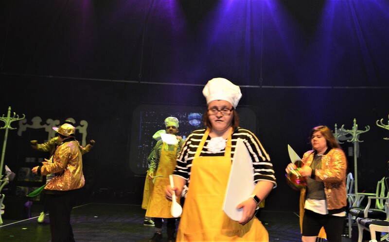 A young female actor in the foreground wearing a whiote bakers hat and yellow apron, holding a two dimensional wooden bowl and wooden spoon props. Other actors in the background in either similar costume of gold sequinned bomber jackets and gold baseball caps. 