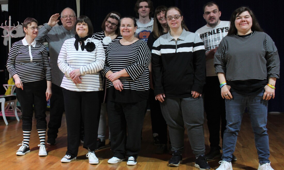 Ten members of Full Circle are pictured in a group format, posing for a photo during a rehearsal for Stomping Ground. They are all smiling and range in ages from 30-77 years old. All are wearing variations on black and white striped costumes.