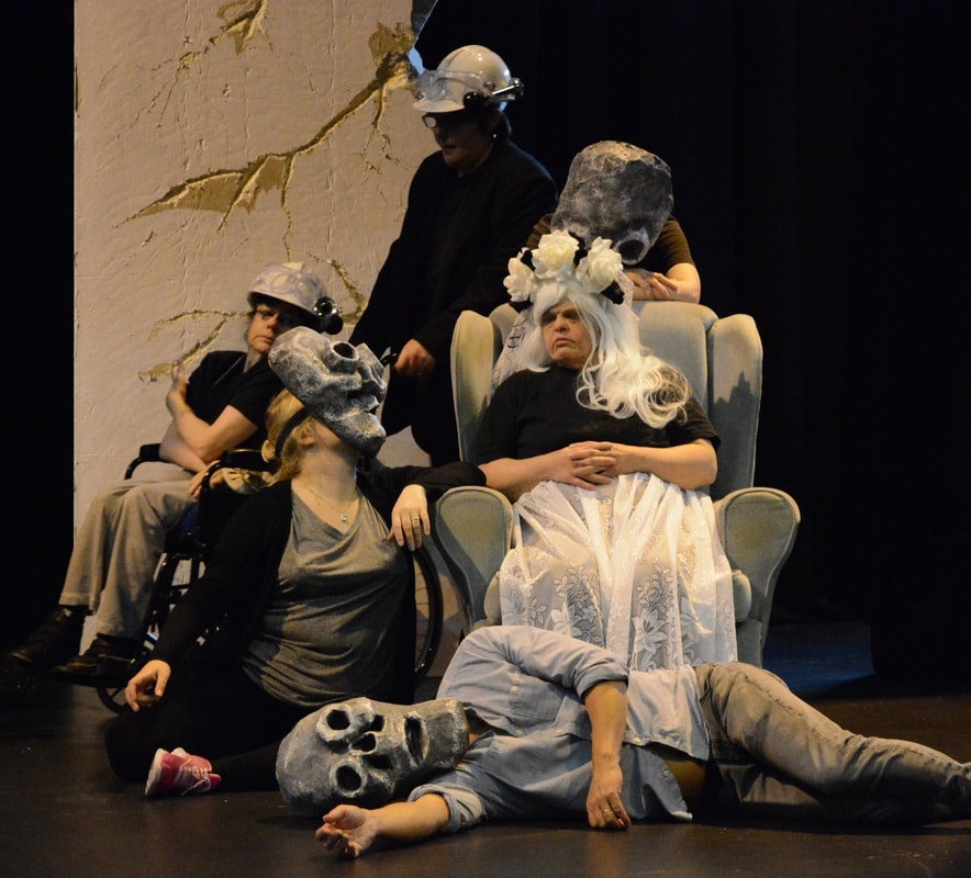 six actors on stage, one is a wheelchair user and another is seated in a large armchair. Everyone is gathered around the armchair. Most actors are wearing large skeleton masks. The seated actor is wearing a stylised white wedding gown and head decoration.