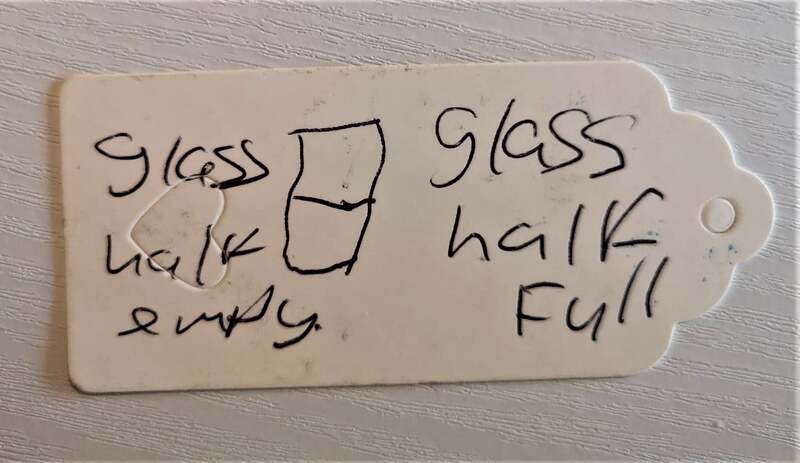 Image Description - White parcel label with handwritten text, 'glass half empty, glass half full' and a line drawing of a half full glass. Pauline Heath.