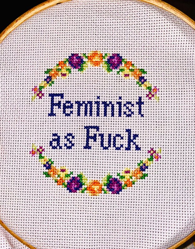 Traditio9nal floral arch at the top and bottom of the cross stitching in orange, purple and yellow with green leaves. Words in the centre read, 'feminist as fuck'.