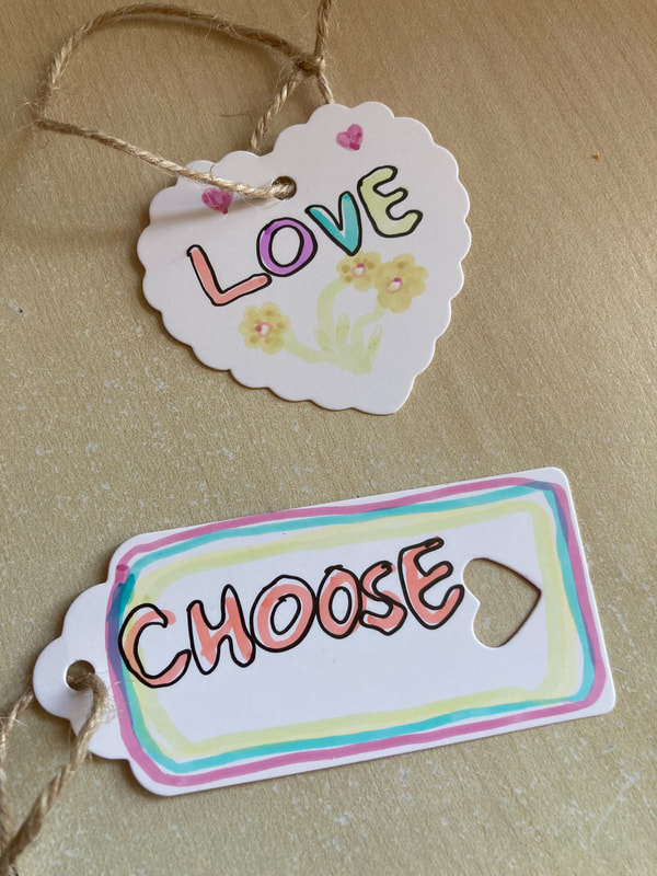 Two labels - one is a white heart with the word love written in pastel colours and three hand drawn yellow flowers. The second says the word choose next to a cut out heart symbolising love. Caroline Miles.