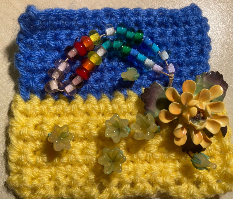 A crocheted Ukrainian flag with a different rainbow of handmade glass beads and an arrangement of different sized glass flowers. Caroline Miles.