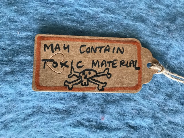 A brown parcel label on a blue wool background. It reads, 'May Contain Toxic Material and had a skull and crossbones on it' Steph Fuller