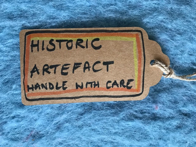 A brown parcel label on a blue wool background. It reads, 'Historic Artefact - Handle with Care' Steph Fuller