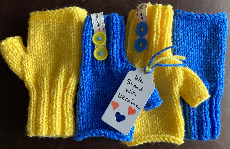 Two pairs of fingerless mittens, alternating blue and yellow, side by side. A lable reading 'we stand with Ukraine' is placed in them with two hand drawn red love hearts.