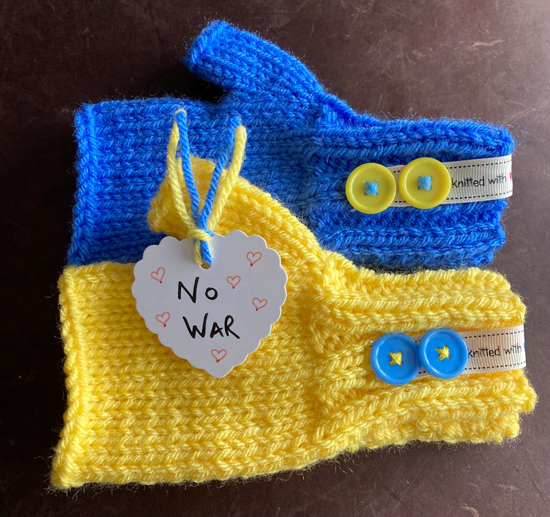 A pair of fingerless mittens, one in blue and one in yellow, both with alternate coloured buttons. A No War love heart label is placed in them.