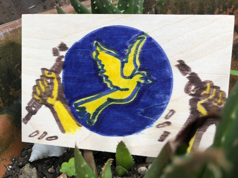 A painted panel with a yellow dove in a blue circle over which two hands are breaking a rifle, bullets falling to the floor. The panel is placed in greenery. JulieMc McNamara.