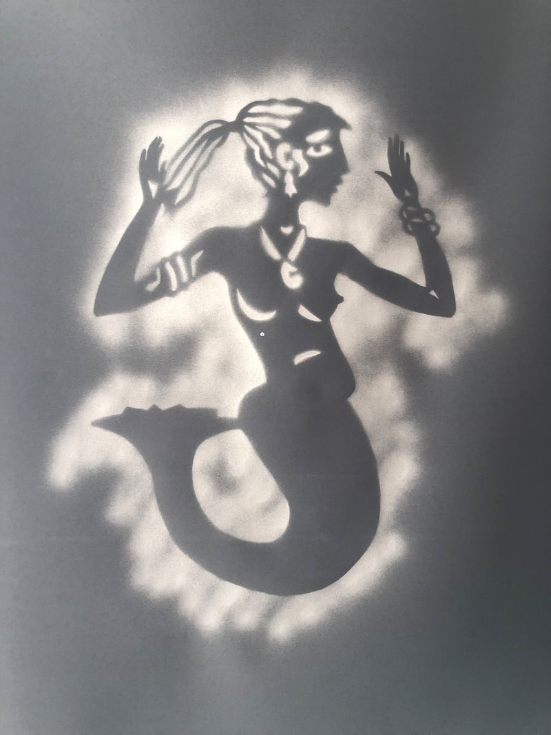Using a handcut stencil of a mermaid facing to the right, there are details of a ponytail,  earring, pendant necklace, bangles, breasts, nipples and buttocks and a mermaid's tail. It had been spray painted white into a charcoal background. 