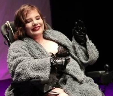 India Robertson is seated smiling in her power chair, performing as a dominatrix. She is a young white woman.