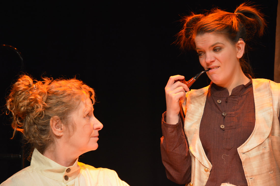 Two white Deaf women actors on stage in Lighthouse by Vici Wreford-Sinnott. They are in brown and cream linen costumes and the younger woman is mock smoking a pipe as she impersonates a man.