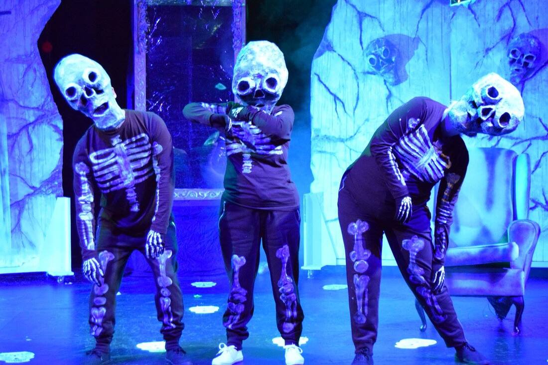 Three actors in quirky poses as skeletons in handpainted costumes and large skeleton head masks, whilst the set in the background shows the run down and spooky walls of a village. 