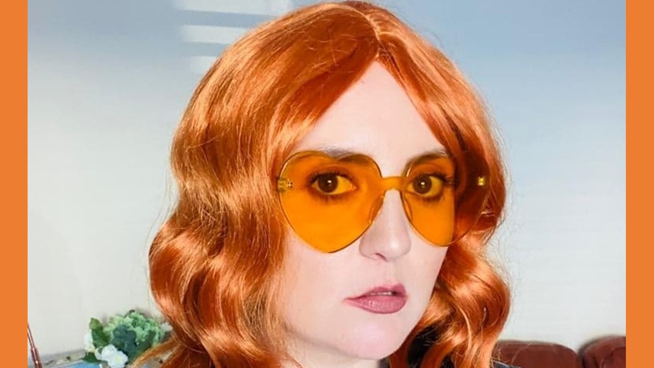 Actor Philippa Cole playing Mim in Siege. She is a young white woman wearing an orange costume wig, large orange love heart glasses and is looking seriously directly into camera. She is wearing a black PVC dress. 