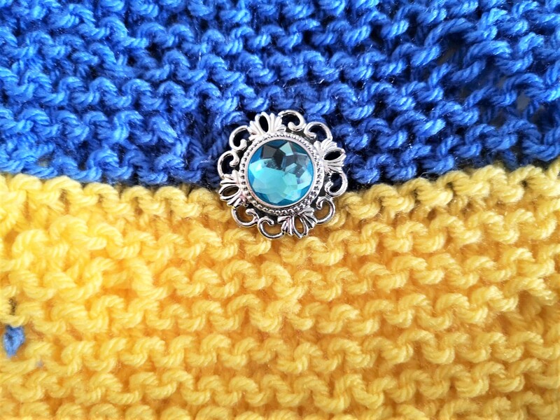Close up of an irregular shaped, hand-knitted Ukrannian flag with a found jewel in the centre. Pale blue with a decorative silver metal surround. Pauline Heath.