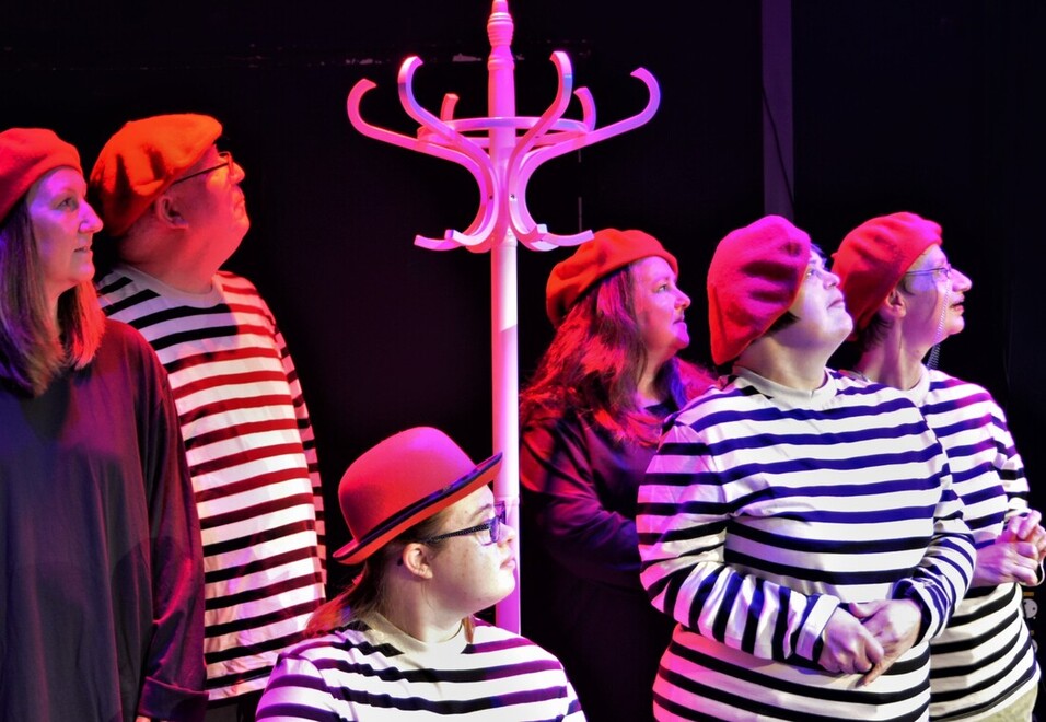 Under red theatre lighting there are six actors. They are gathered around a white wooden hat stand. They are wearing a mix of bold black and white striped tops and plain black tops. They are all wearing hats, five are wearing red berets and one is wearing a red bowler hat. The actors are all looking off into the distance.
