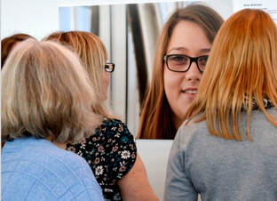 There are the backs of three women's heads, looking at a large, photographed portrait of a young women with long brown hair, who is wearing glasses. In the portrait she is smiling.