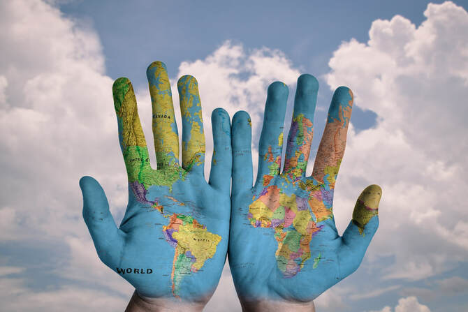 Two hands being held up against a blue sky with white clouds. The hands have palms facing us and are painted with a map of the world.