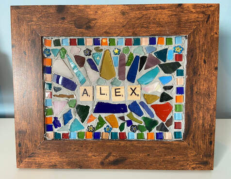 Mosaic by Alex Dechbamrung. The mosaic kits were created by Little Cog inspired by work seen on the internet. Within a brown wooden picture frame is a mosaic of each participant's name, created using scrabble style letter tiles, in this case Alex. The mosaic is created from a number of small square glass tiles in a range of colours and broken pieces of glass, again in a range of colours, arranged decoratively and grouted in position. Some strong blues, oranges and greens mixed in.