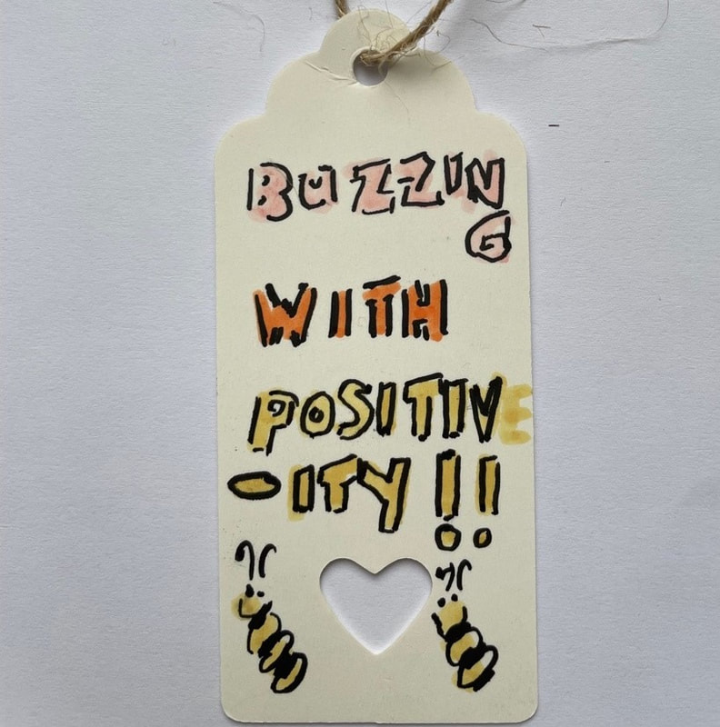 A white parcel label with a heart shape cut out. Two little bees are drawn and it reads Buzzing with positive-ity. Samantha Blackburn