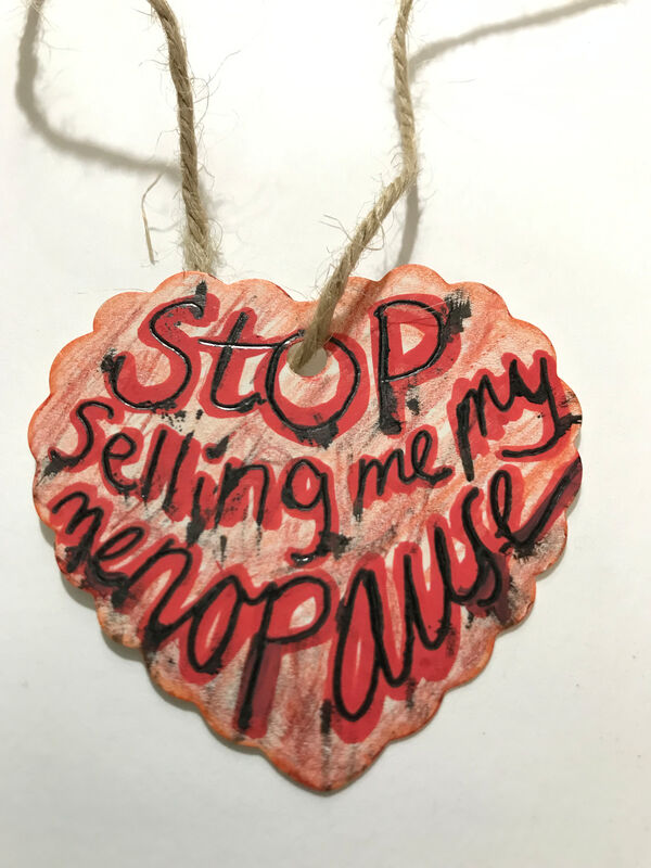 A heart shaped label with a background colour of scratchy pink. Bold black and red lettering says 'stop selling me my menopause' Caroline Cardus