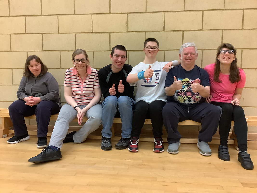 a group of six very happy learning disabled people seated on a low sports bench in a sports hall. Three of them have their thumbs up and all are wearing sports shoes ready for action. 