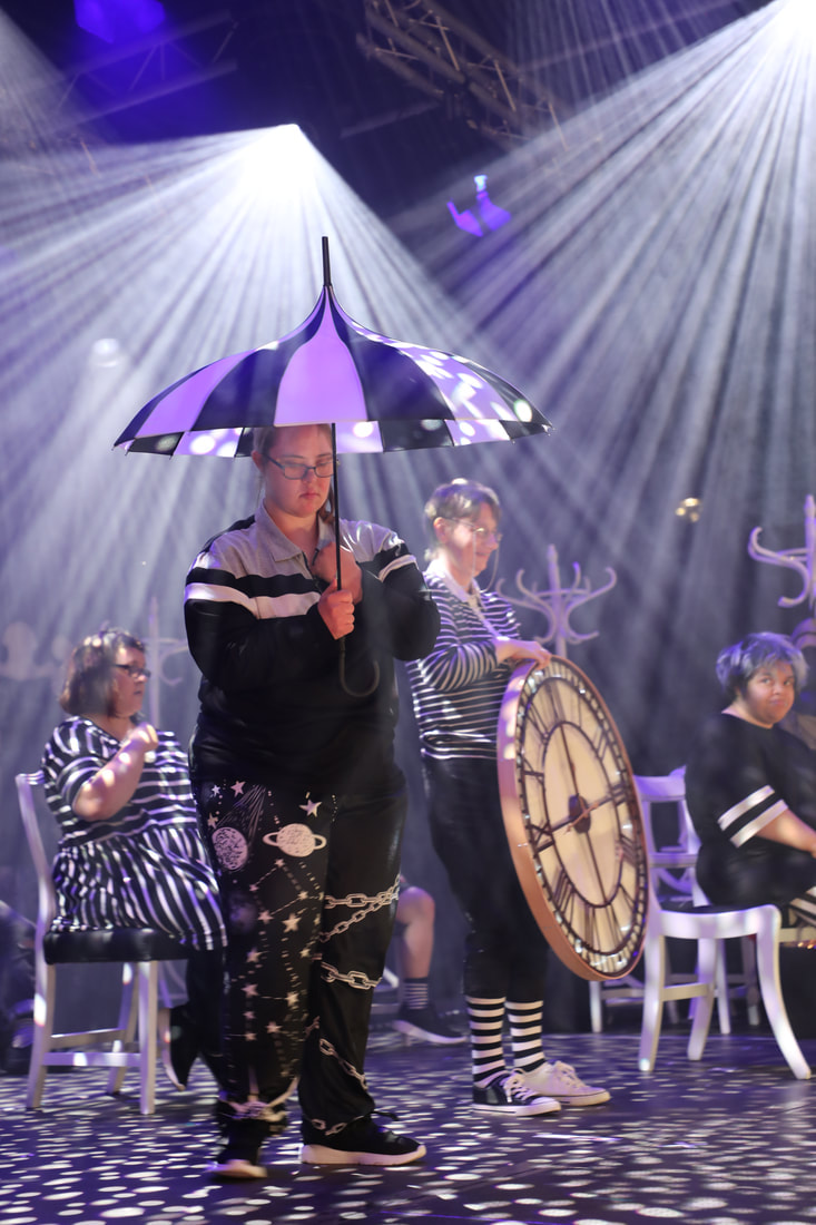 Another image from the live SIRF show, actors in a bold lighting effect of downward shafts of light. One actor is holding a black and white pagoda umbrella, another is holding a giant clock, whilst others are sitting on chairs trying to save their memories from disappearing.