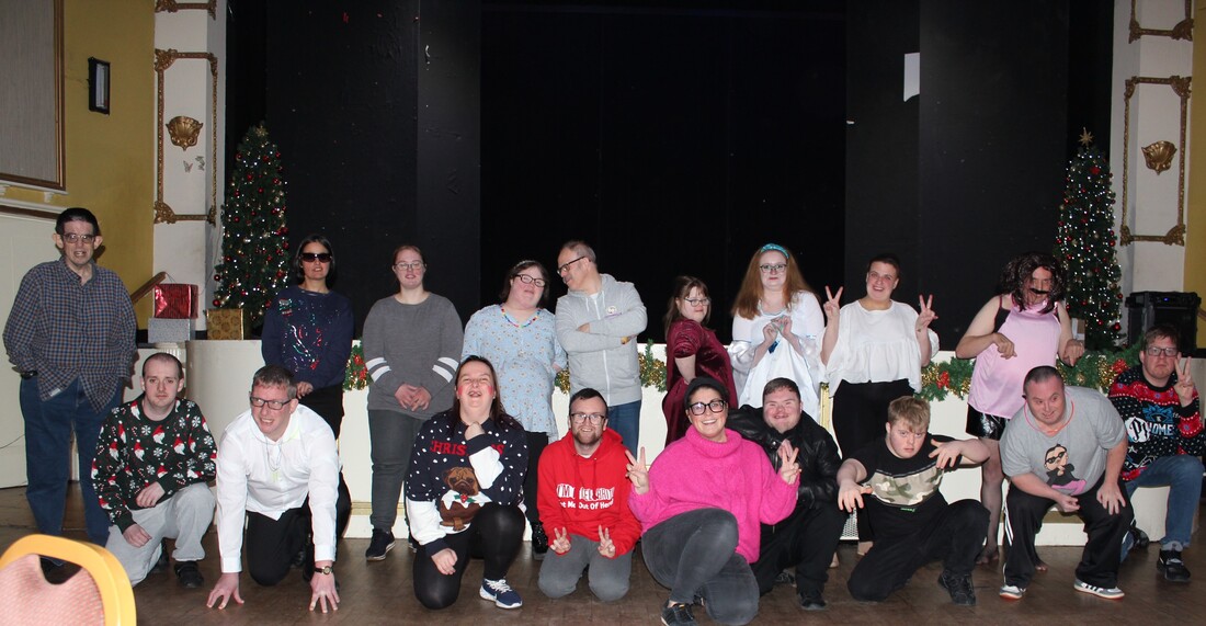 18 learning disabled actors al posing as a group in front of a stage. The front row are all seated on the floor while others stand behind them. All are smiling and several are making the peace sign with their hands. It's a photo taken at Christmas time so some people are in Christmas jumpers and there are Christmas decorations around the room. 