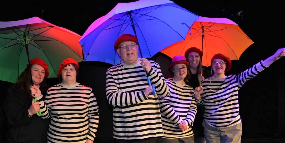 Six happy actors standing underneath colourful umbrellas. All are wearing a mix of plain black tops and stripey black and white tops, red berets and one person is wearing a red bowler hat. They are laughing and cheering and one person has her arm raised in celebration. 